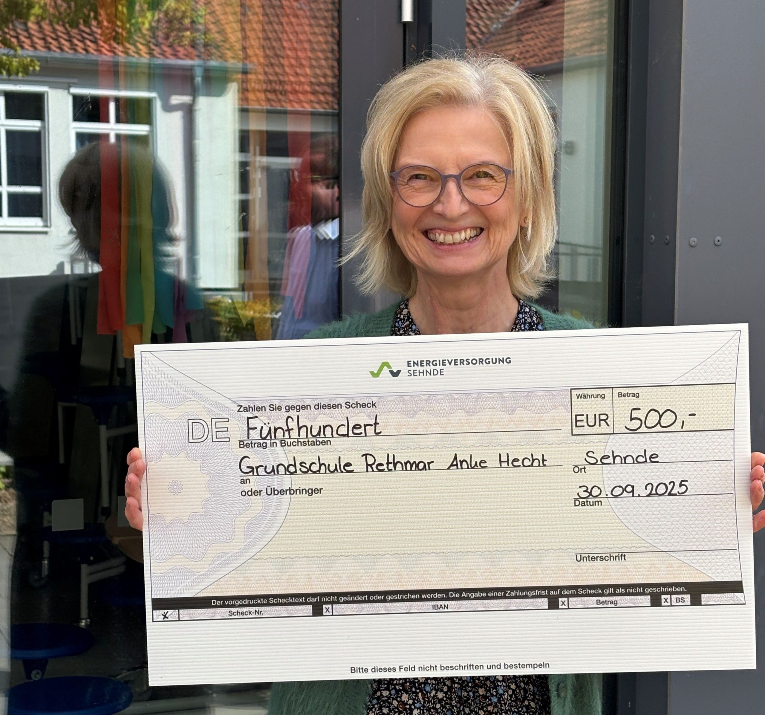 Person hält einen großen symbolischen Scheck vor einem Gebäude mit Glasfront. Der Scheck zeigt: ‚Energieversorgung Sehnde‘, Betrag 500 €, Empfänger Grundschule Rethmar