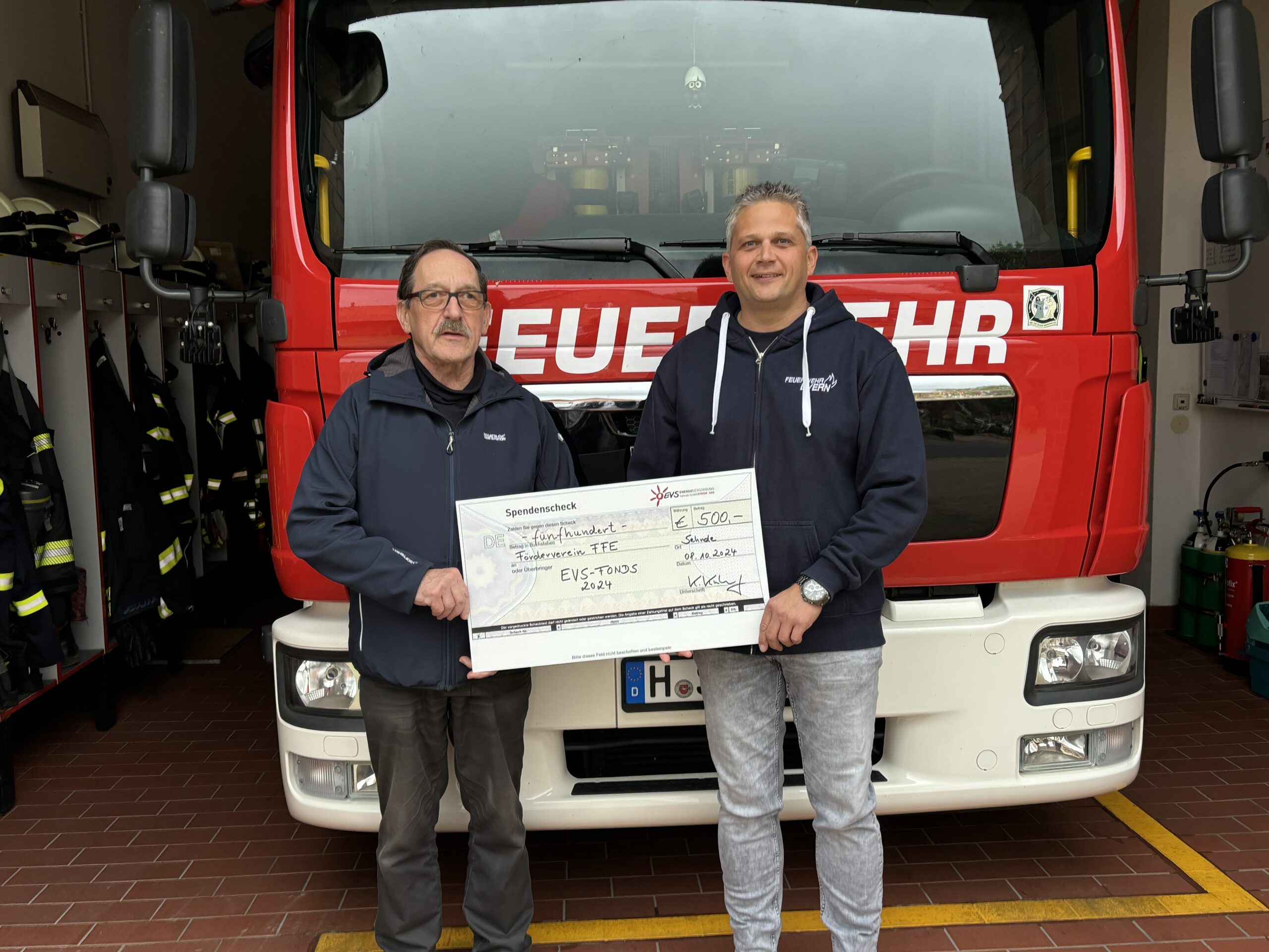 Scheckübergabe im Rahmen des EVS-Fonds 2024, Heinrich Klinge (Vorsitzender des Fördervereins der FFW Evern) und Matthias Söchtig (Ortsbrandmeister) halten den Spendenscheck in den Händen und stehen vor einem Einsatzfahrzeug