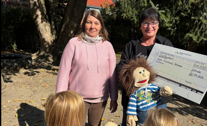 Zwei Personen im Freien mit großem Scheck über 500 € für Kita Huckmuck, Rethmar, Kinder und Handpuppe im Vordergrund.