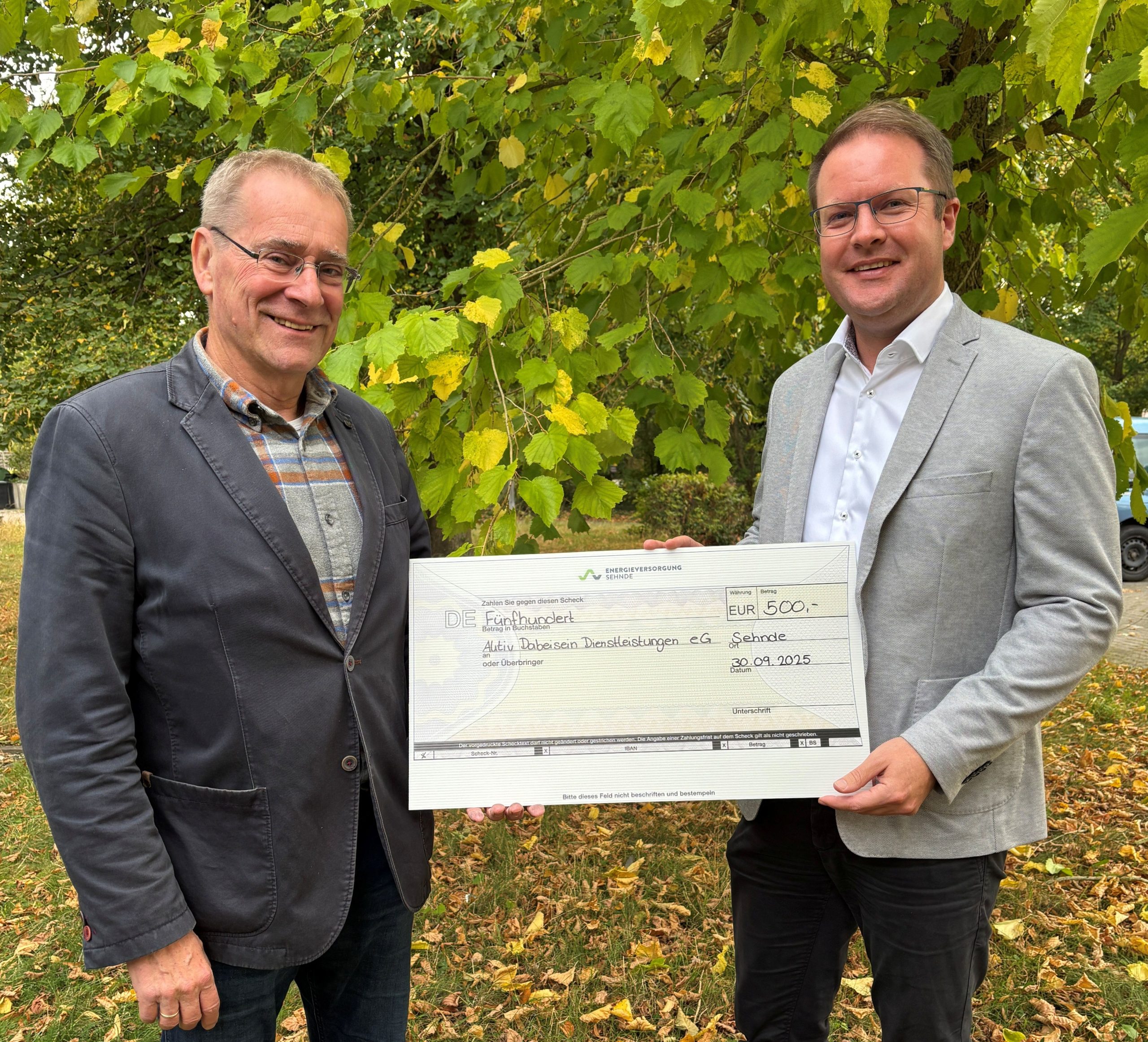 Zwei Personen im Freien halten einen großen symbolischen Scheck über 500 € für die Aktiv Dabeisein Dienstleistungen eG, datiert 30.09.2025.