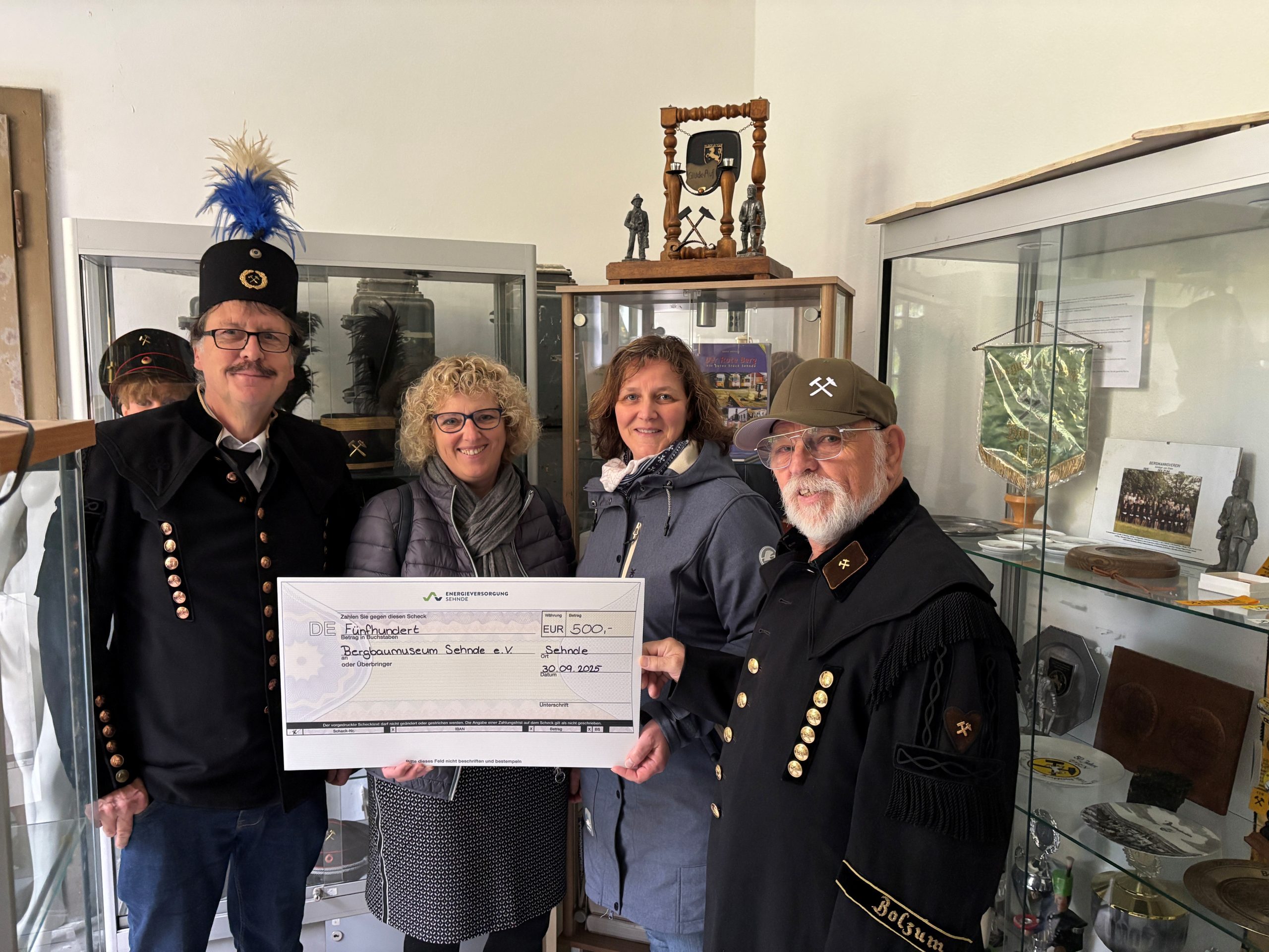 Vier Personen stehen in einem Raum mit Vitrinen voller Bergbau-Exponate und halten gemeinsam einen großen symbolischen Scheck über 500 Euro für den Bergbauverein Sehnde e.V.