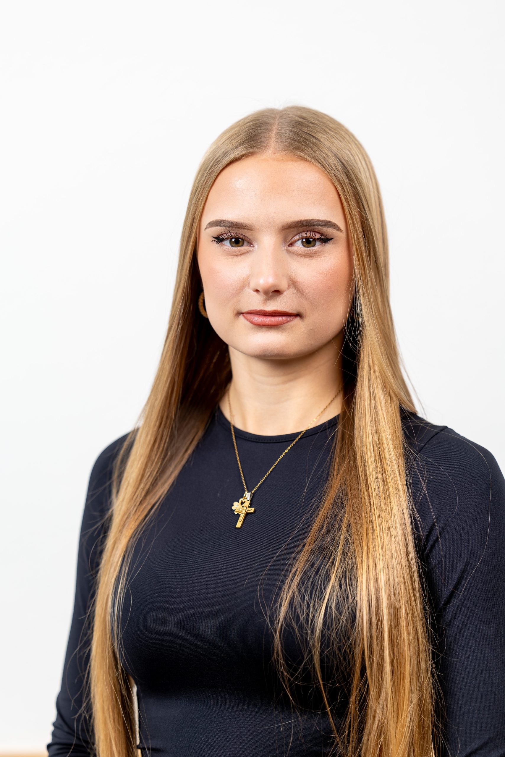 Person mit sehr langen, glatten, hellbraunen Haaren, trägt ein schwarzes, langärmliges Oberteil und eine goldene Halskette mit Kreuzanhänger. Hintergrund ist schlicht weiß