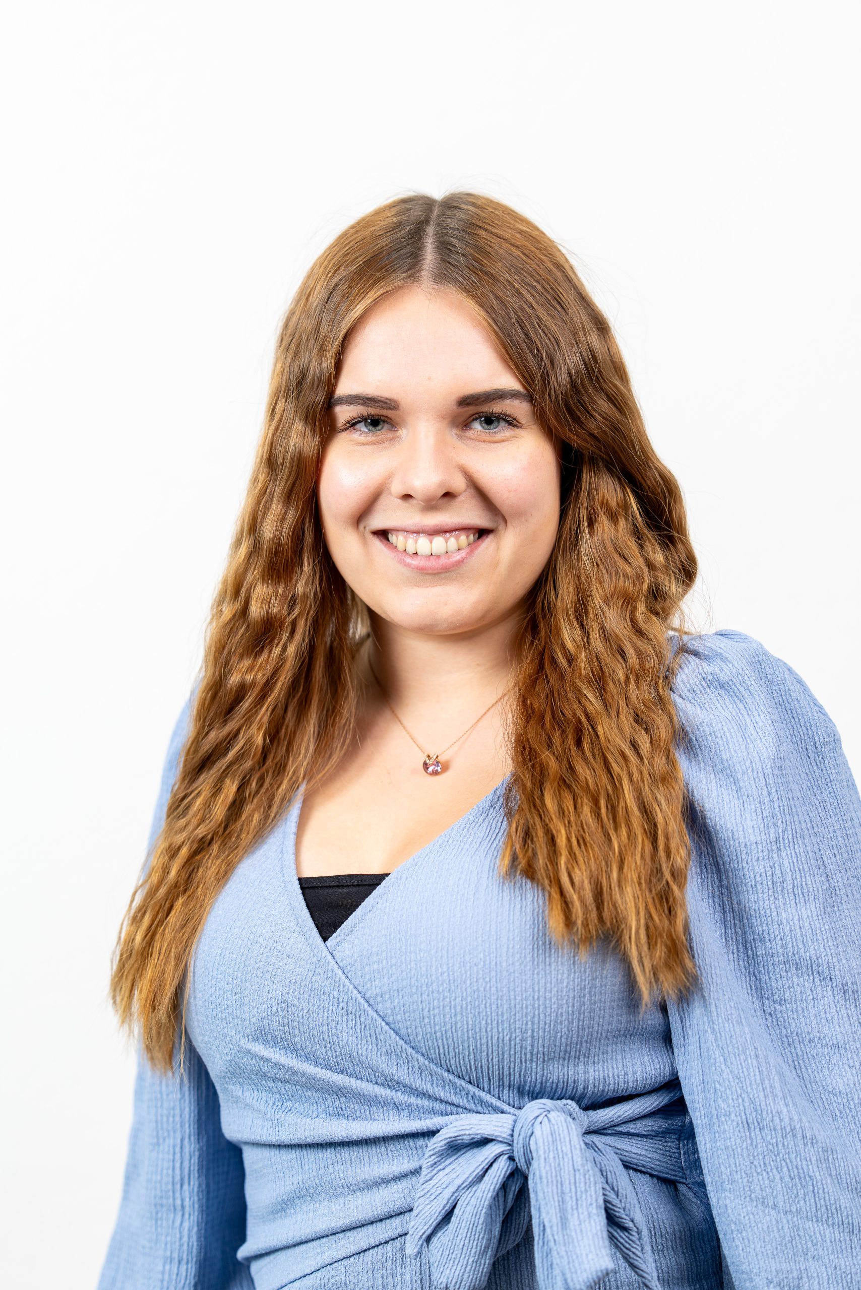 Person mit langen, gewellten, hellbraunen Haaren, trägt eine hellblaue Wickelbluse mit Bindeschleife und einen dezenten Halskettenanhänger. Hintergrund ist einfarbig weiß