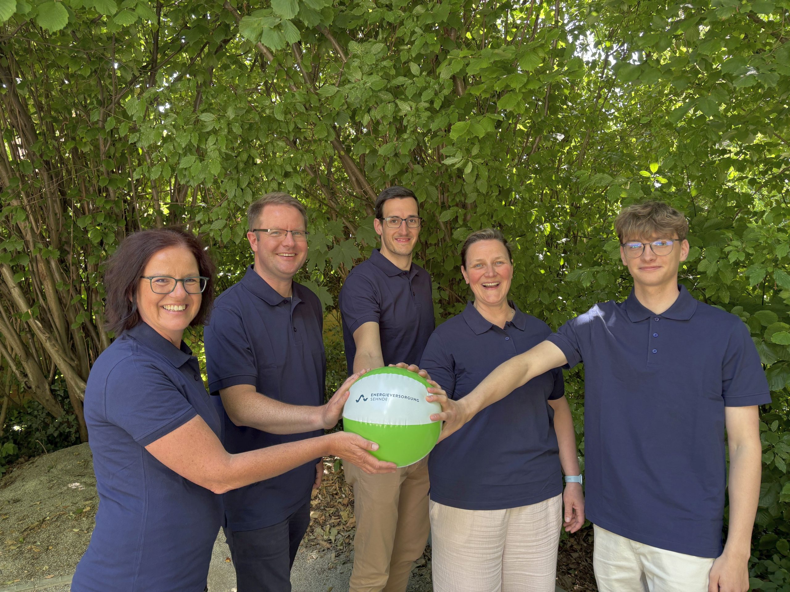 Fünf Personen in blauen Poloshirts stehen vor grünem Laub und halten gemeinsam einen grünen und weißen Ball mit dem Logo ‚Energieversorgung Sehnde‘