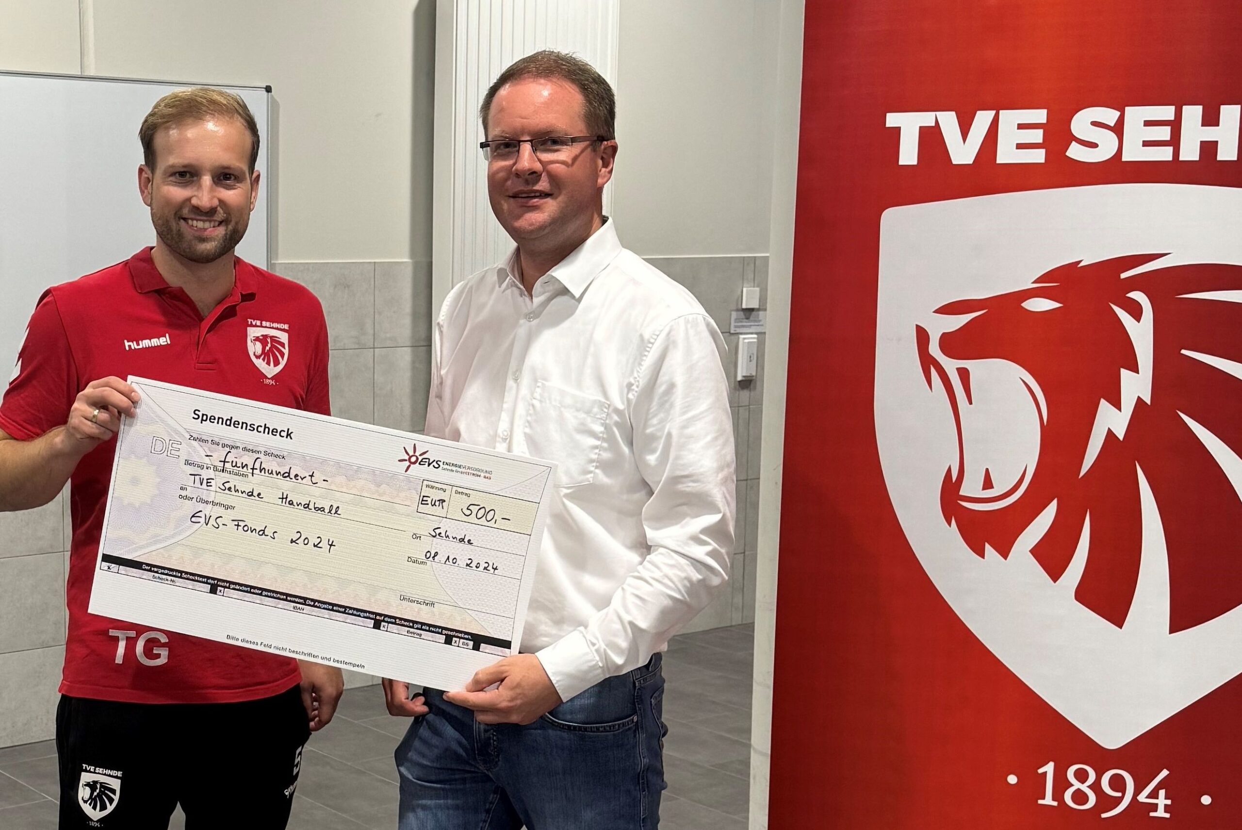 Scheckübergabe im Rahmen des EVS-Fonds 2024, Timo Gewohn (TVE Sehnde und Timo Rüffer (Geschäftsführer EVS) stehen neben einem Roll-Up des TVE in der Sporthalle Chausseestraße in Sehnde und halten den Spendenscheck über 500 Euro in den Händen.